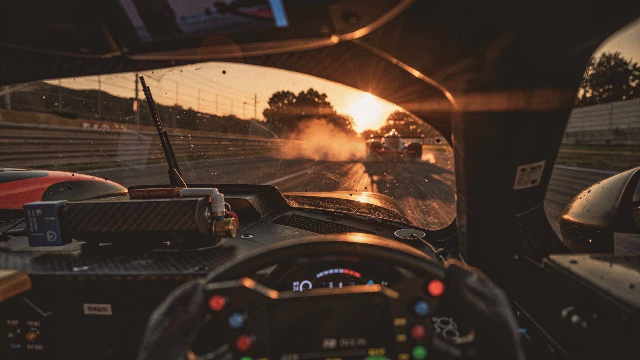 Driver's-eye view from a race car cockpit at dusk, chasing a rival car through a cloud of dust into the low sun.